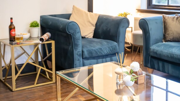 A modern living room with a blue sofa, a glass coffee table, and a small side table with a bottle and glasses. - Northern Hideaways, Apple Store, Ellingham (Chathill)