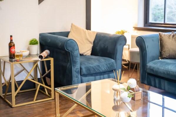 Ein modernes Wohnzimmer mit einem blauen Sofa, einem Glastisch und einem kleinen Beistelltisch mit Flasche und Gläsern.