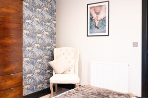 Ein Zimmer mit ornamentalem Wandpapier, einem gerahmten Kunstwerk an der Wand, einem gepolsterten Sessel und einer Heizung.
