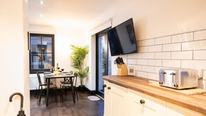 A modern kitchen with white cabinets, a wooden countertop, a flat-screen TV mounted above the counter, and a dining area with a table and chairs. - Northern Hideaways, Apple Store, Ellingham (Chathill)
