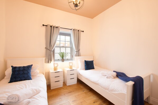 A bedroom with two single beds, bedside tables, a window with curtains, a potted plant, and a ceiling light fixture.