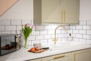 A kitchen with white tiles, a gold faucet, a toaster, a bowl, a pomegranate, and a vase with flowers. - Northern Hideaways Eider Cottage (Seahouses)