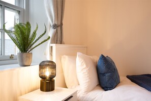 A neatly made bed with white and blue pillows, a bedside lamp, and a potted fern on a bedside table. - Northern Hideaways Eider Cottage (Seahouses)