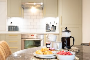 A kitchen with a marble countertop, a coffee maker, a plate of croissants, a bowl of fruit, and a white mug. - Northern Hideaways Eider Cottage (Seahouses)