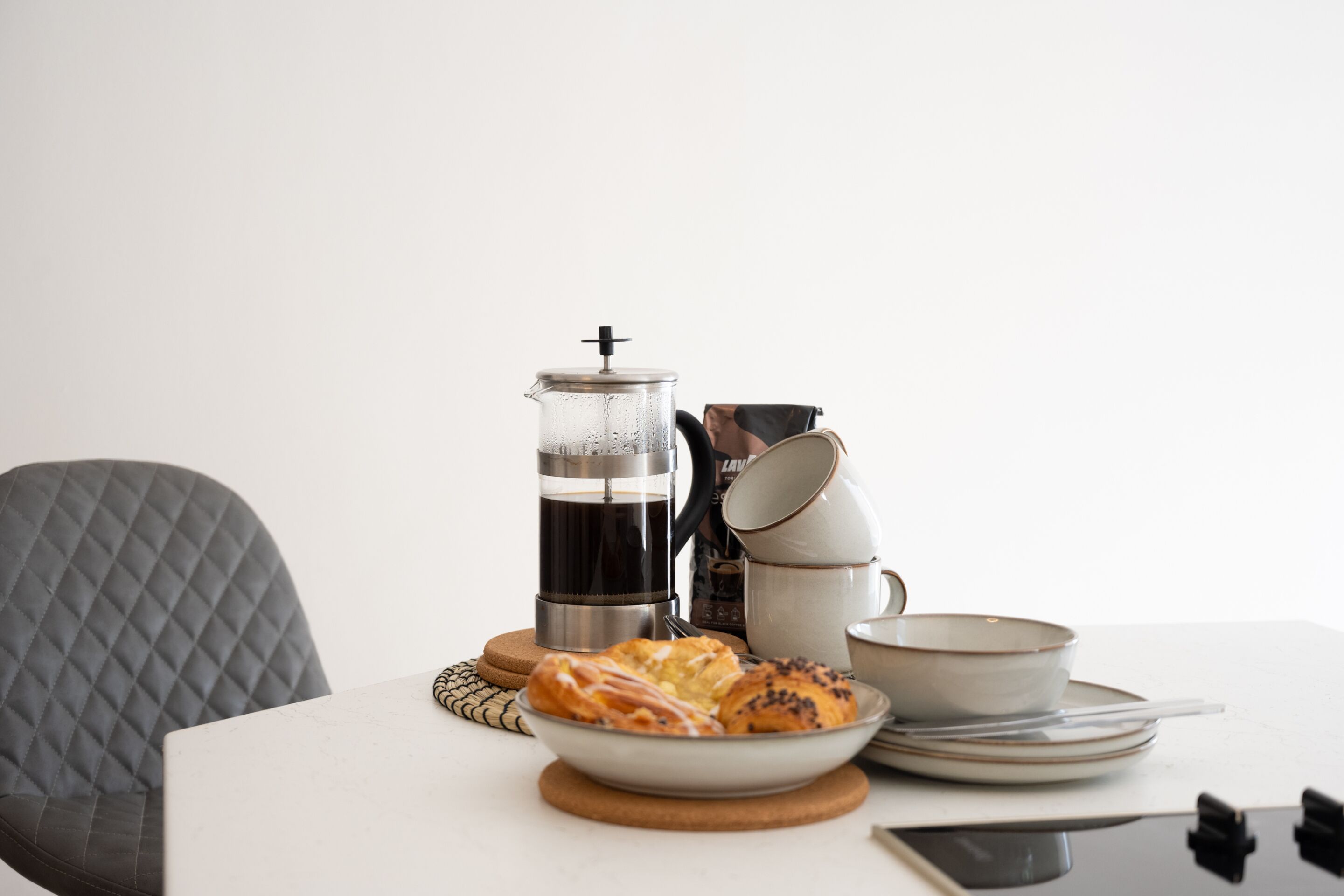 A table with a coffee maker, a stack of cups, a bowl of pastries, and a smartphone.
