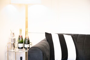 Un sofá negro con un cojín a rayas blancas y negras, una mesa de cristal con dos botellas de champán, y una lámpara al fondo.