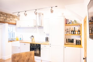 A modern kitchen with white cabinets, a wooden countertop, and a built-in oven. There is a bookshelf with books and bottles, and a stone wall in the background.