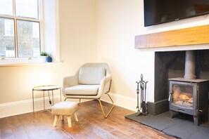 A living room with a fireplace, a comfortable armchair, a small table, and a television mounted on the wall. - Northern Hideaways, St Cuthberts (Seahouses)