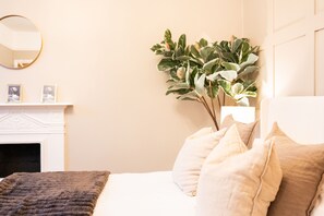 Un dormitorio con cama, almohadas, un espejo, chimenea y una planta.