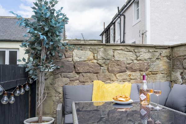 Una mesa de vidrio con una botella de vino, dos copas y un plato de comida en un entorno al aire libre con paredes de piedra y una valla de madera.