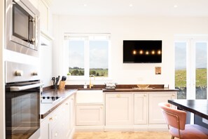 Une cuisine moderne avec des meubles blancs, un four encastré et une grande fenêtre donnant sur la campagne.