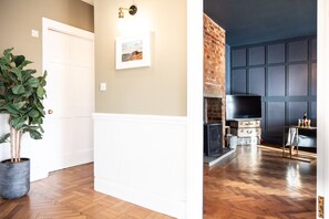 Une pièce avec un plancher en bois, une porte blanche, une plante en pot, un tableau encadré accroché au mur, et une cheminée en briques sur laquelle sont posés un téléviseur et des boîtes de rangement.