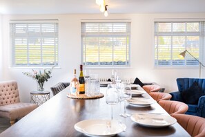 A dining area with a long table set for a meal, two windows with blinds, a vase with flowers, and a bottle of wine.