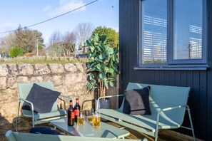 A patio with two green chairs, a table with bottles and glasses, and a view of a stone wall and trees.