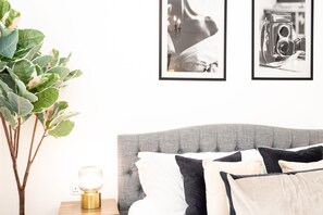 A bedroom with a grey upholstered headboard, white and black pillows, a bedside lamp, and a potted plant. Two black and white photographs are displayed on the wall.