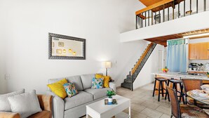 A modern living room with a grey sofa, a white coffee table, and a dining area with wicker chairs. A staircase leads to an upper level.