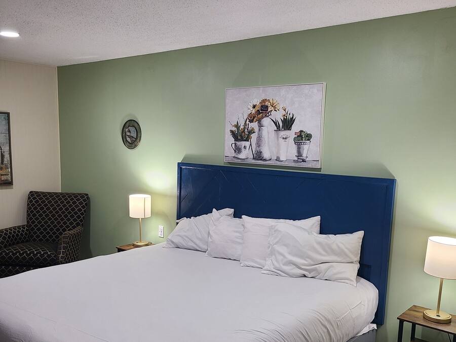 A neatly made bed with white linens, a blue headboard, and a wall-mounted painting of potted plants.