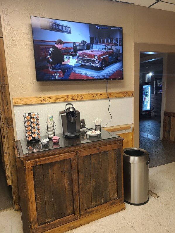 Un coin bar avec un téléviseur fixé au mur diffusant une émission sur les voitures, une machine à café et une poubelle.
