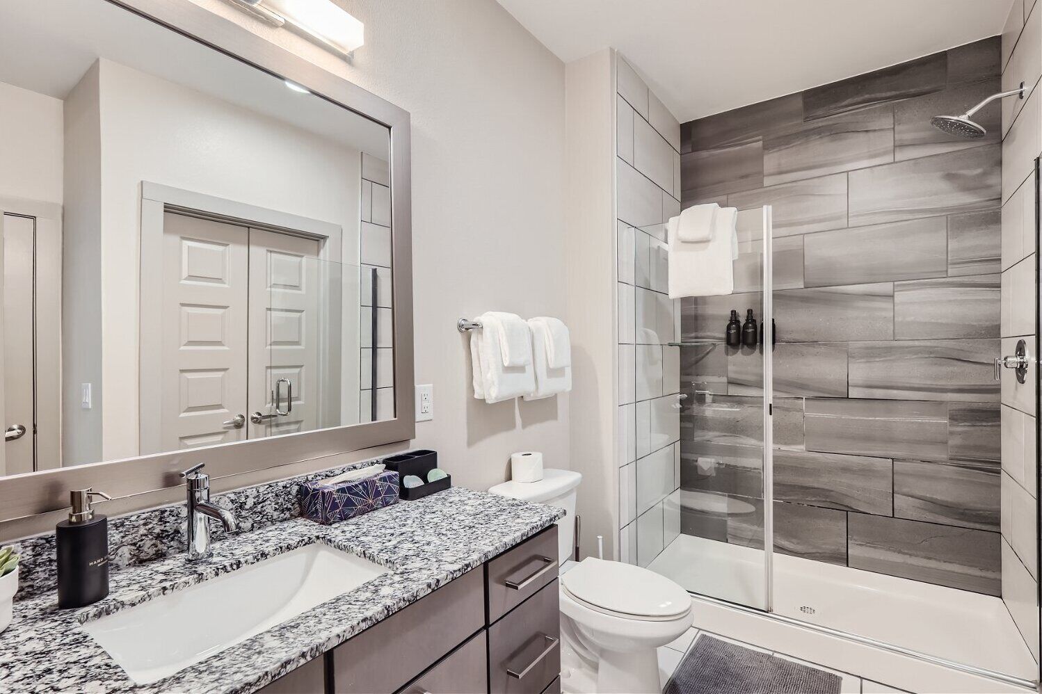 Une salle de bain moderne avec un comptoir en granit, un grand miroir, une douche avec une porte en verre et des toilettes.