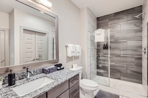 Un baño moderno con encimera de granito, un espejo grande, ducha con puerta de vidrio y un inodoro.
