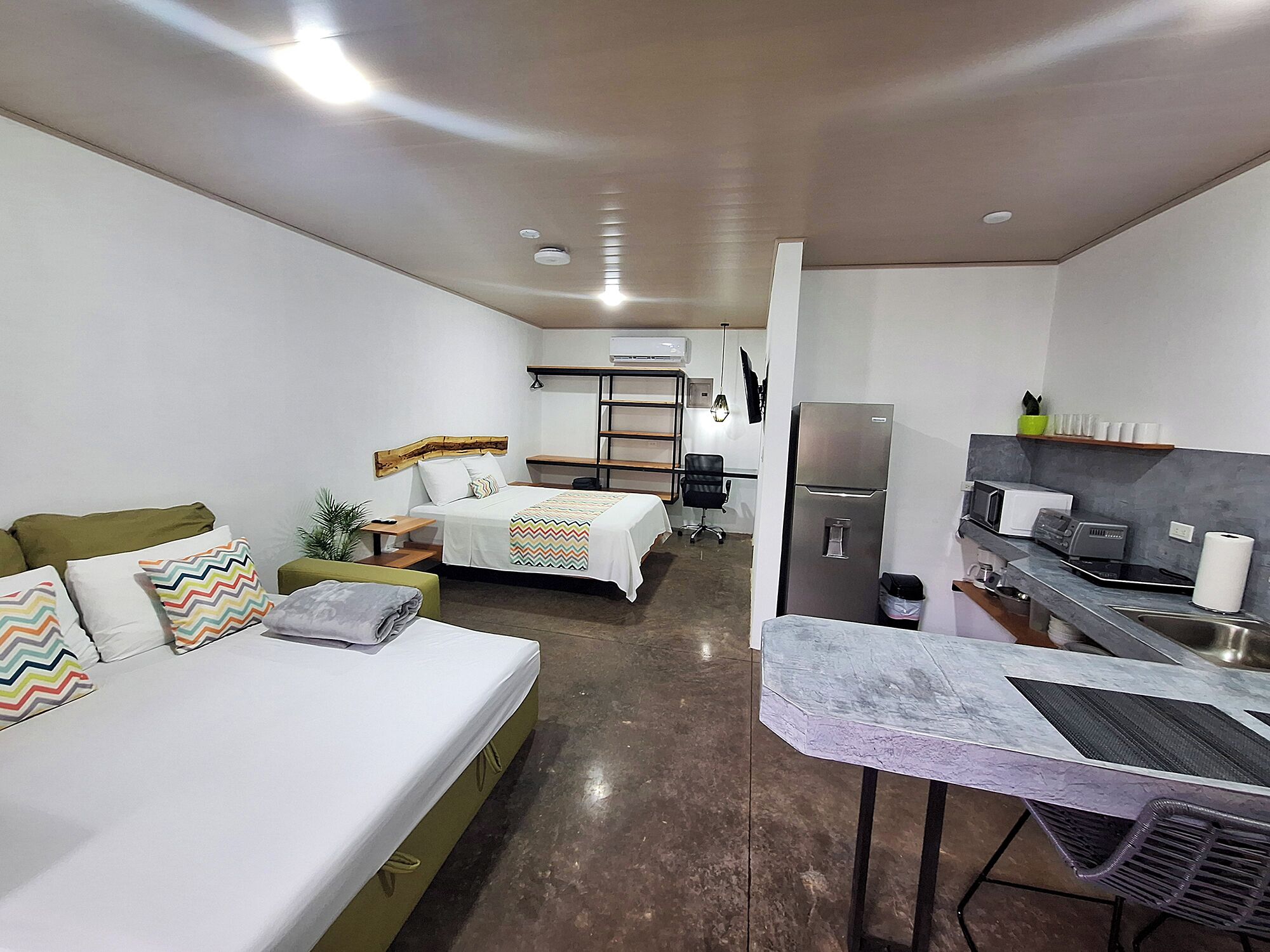 A compact hotel room with a bed, a small desk, a chair, a kitchenette, and a bathroom.