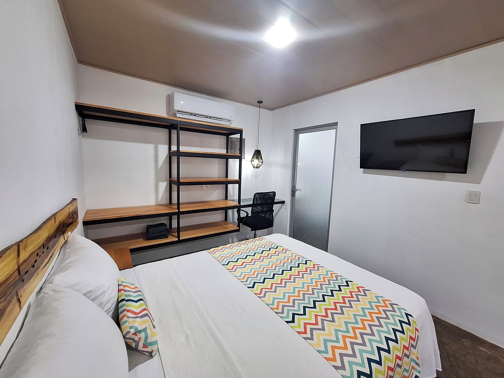 A bedroom with a bed, a TV mounted on the wall, a wardrobe, and a desk with a chair.