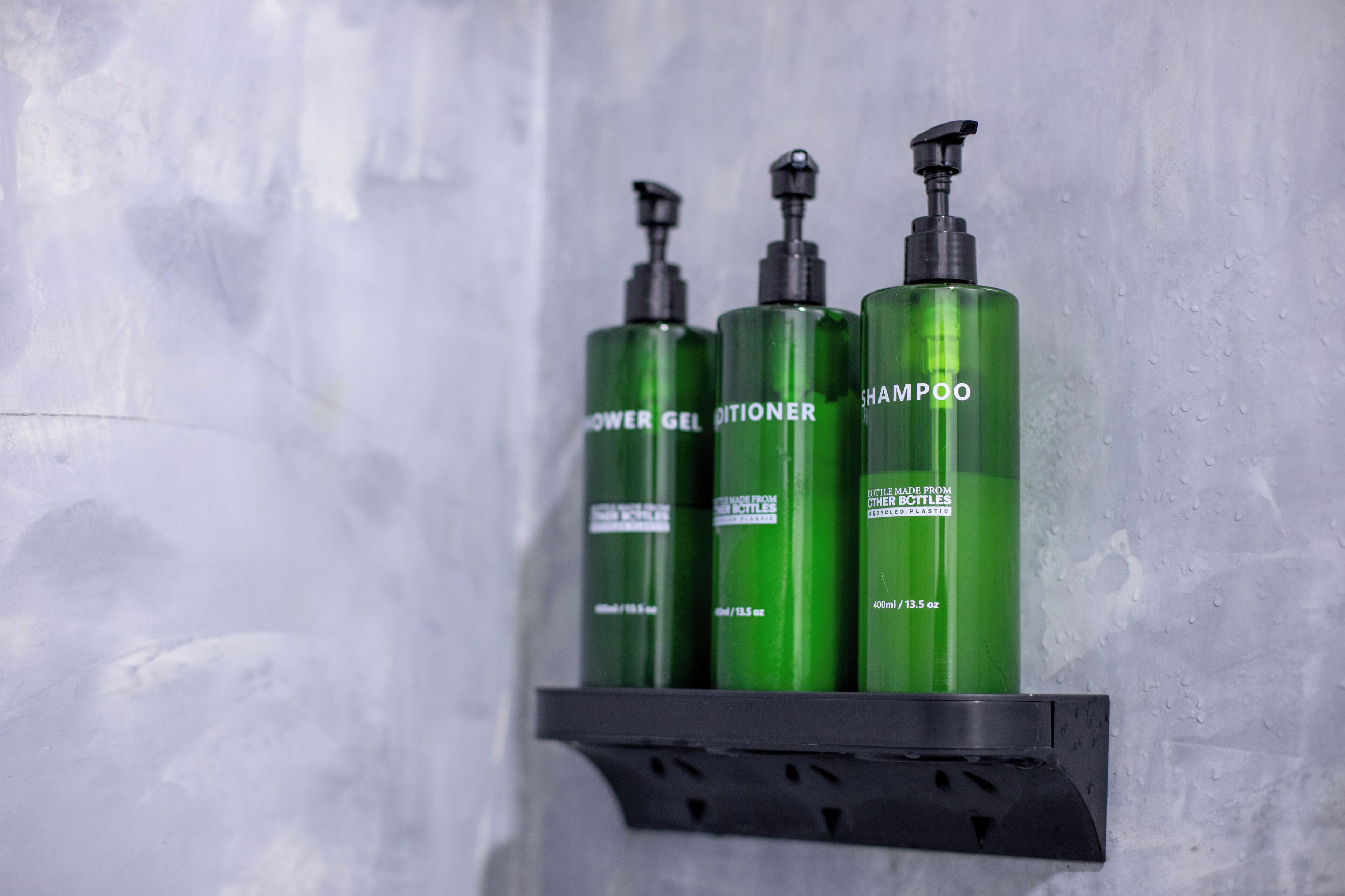 Three green bottles of shampoo, conditioner, and shower gel on a black shelf against a textured wall.