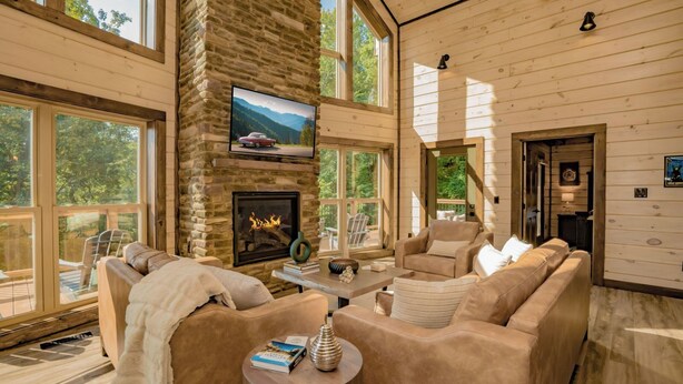 A cozy cabin interior with a stone fireplace, comfortable seating, and large windows offering a view of the outdoors. - Smoky Mountain Skies - Mountain views, gorgeous interiors, game room, hot tub, and more! (Gatlinburg)