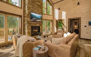A cozy cabin interior with a stone fireplace, comfortable seating, and large windows offering a view of the outdoors. - Smoky Mountain Skies - Mountain views, gorgeous interiors, game room, hot tub, and more! (Gatlinburg)