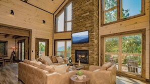 A spacious living room with a stone fireplace, large windows, and a television. - Smoky Mountain Skies - Mountain views, gorgeous interiors, game room, hot tub, and more! (Gatlinburg)