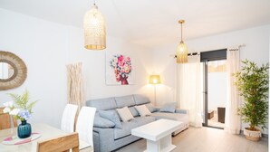 A modern living room with a grey sofa, white coffee table, and a dining area with a glass table and wooden chairs. - Casa Flor del Rocío – Seville – 4 Bedrooms for 8 – AC Throughout – Terrace & Parking (La Rinconada)