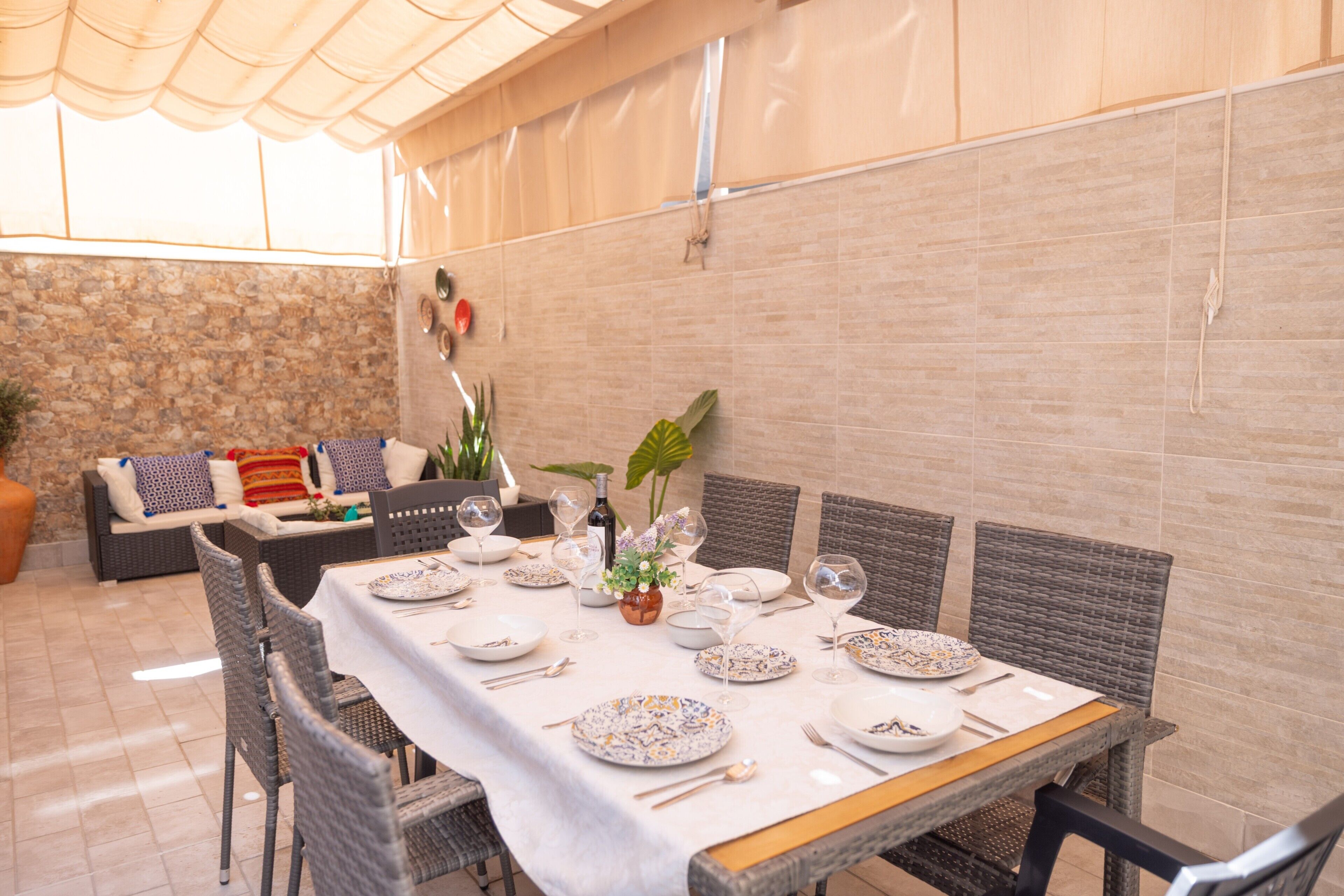 Een eetruimte met een tafel gedekt voor zes personen, omringd door rieten stoelen, een stenen muur en een plafond met een lamp.