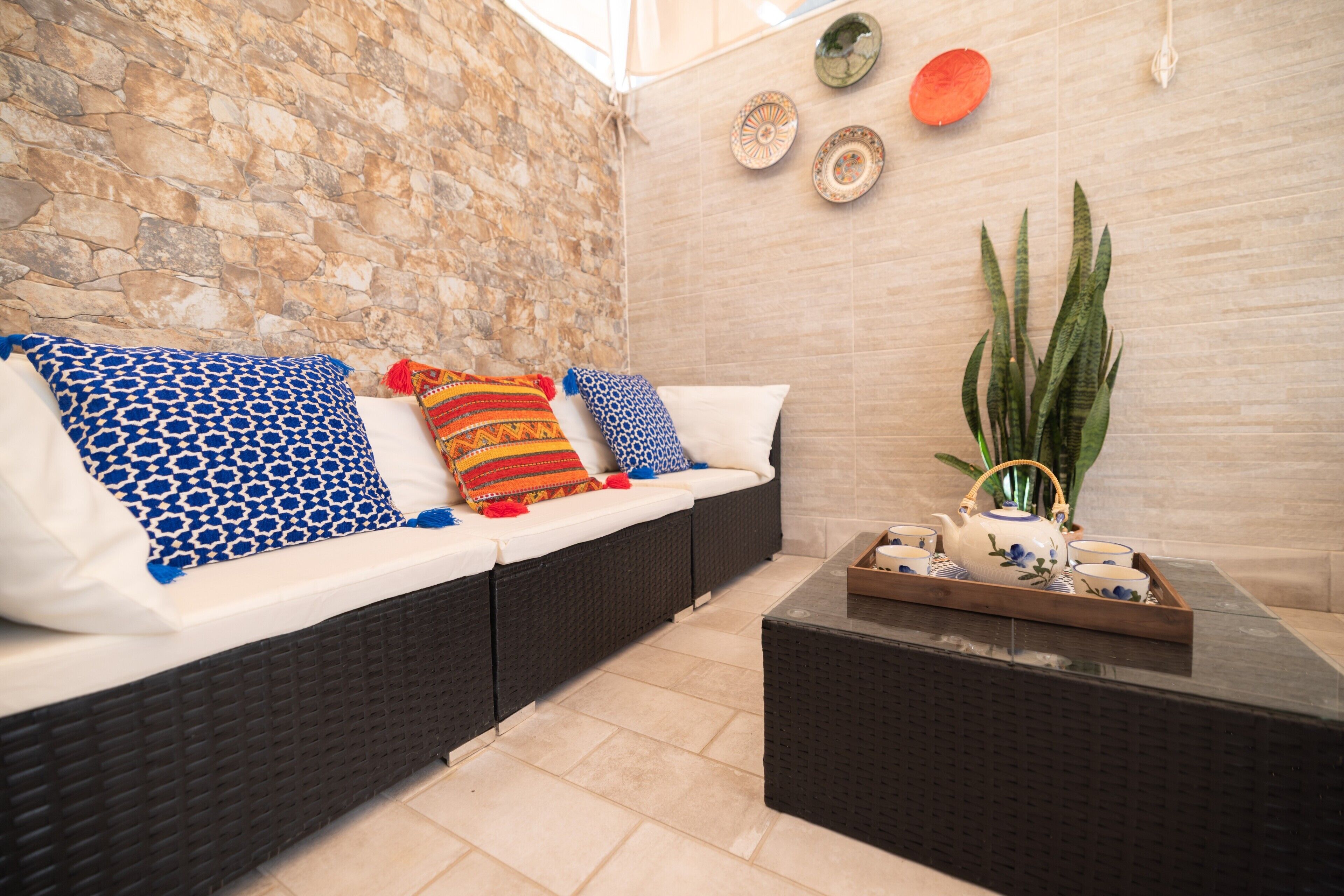 A room with a stone wall, a sofa with patterned cushions, a tray with teapot and cups, and a large plant.