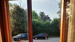 Une vue à travers une fenêtre avec un cadre en bois, une rambarde en métal rouge et une zone pavée où sont garées des voitures.
