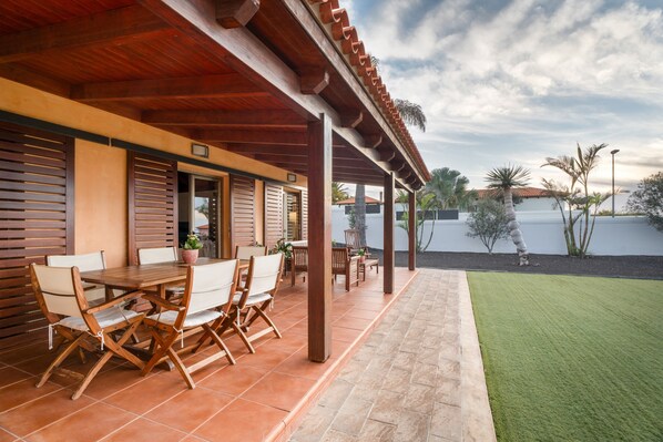 An outdoor patio with a wooden table and chairs, a potted plant, and a view of a building and palm trees.