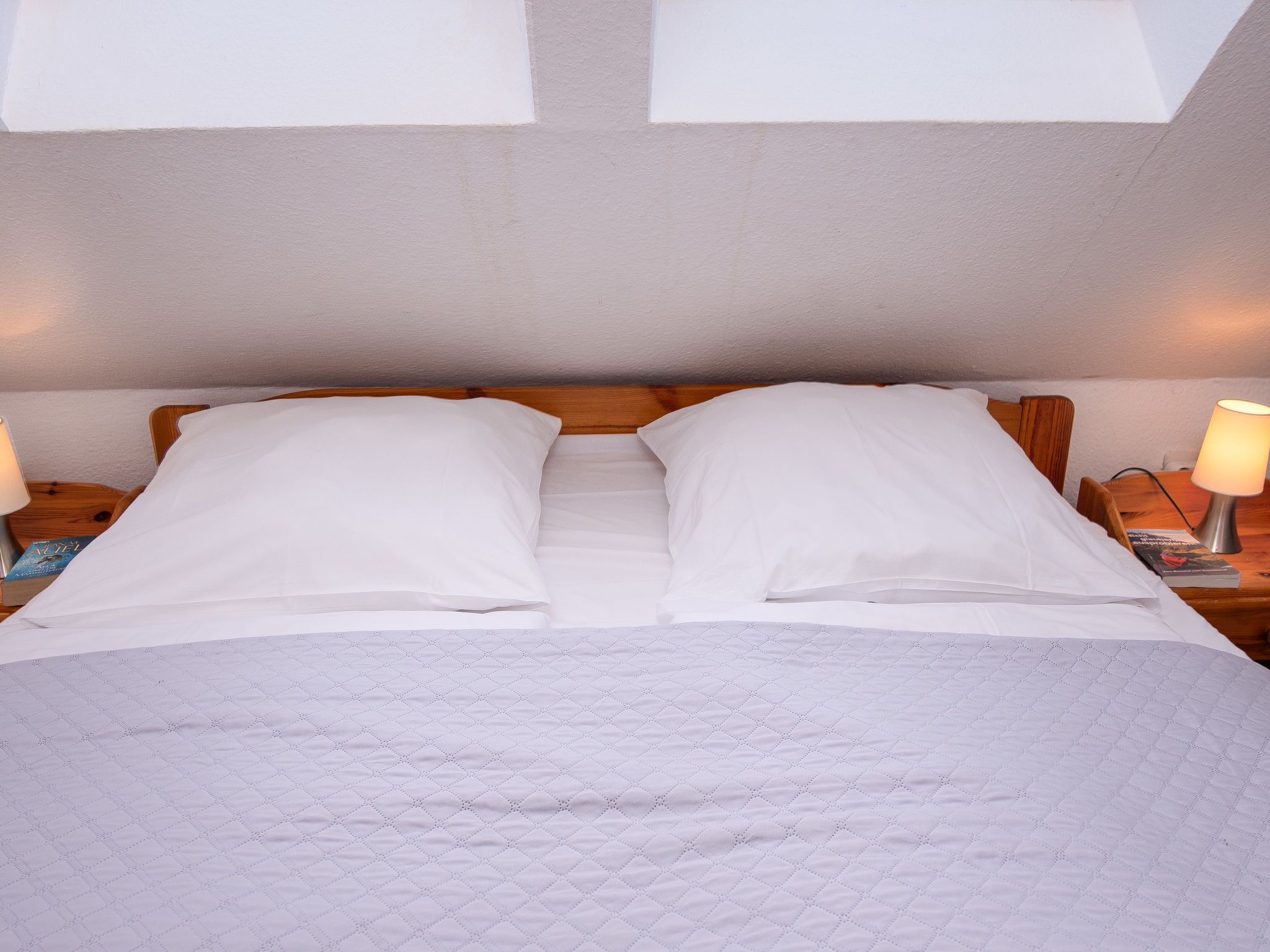 Ein ordentlich gemachtes Bett mit weißen Leinen und einer gesteppten Decke, zwei Nachttischlampen und einem Holz-Kopfteil.