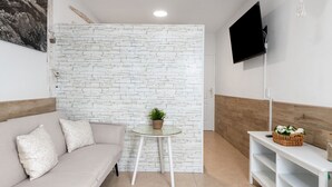 Quarto moderno com parede de tijolos brancos, sofá com almofadas estampadas, mesa redonda pequena com uma planta, TV fixada na parede e cabeceira de madeira.