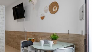Uma sala de jantar com uma mesa redonda, duas cadeiras, uma televisão fixada na parede e um vaso de planta.