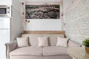 A neutral-toned sofa with patterned cushions, a wall-mounted picture of a landscape, a white brick wall, and a small round table with a plant.