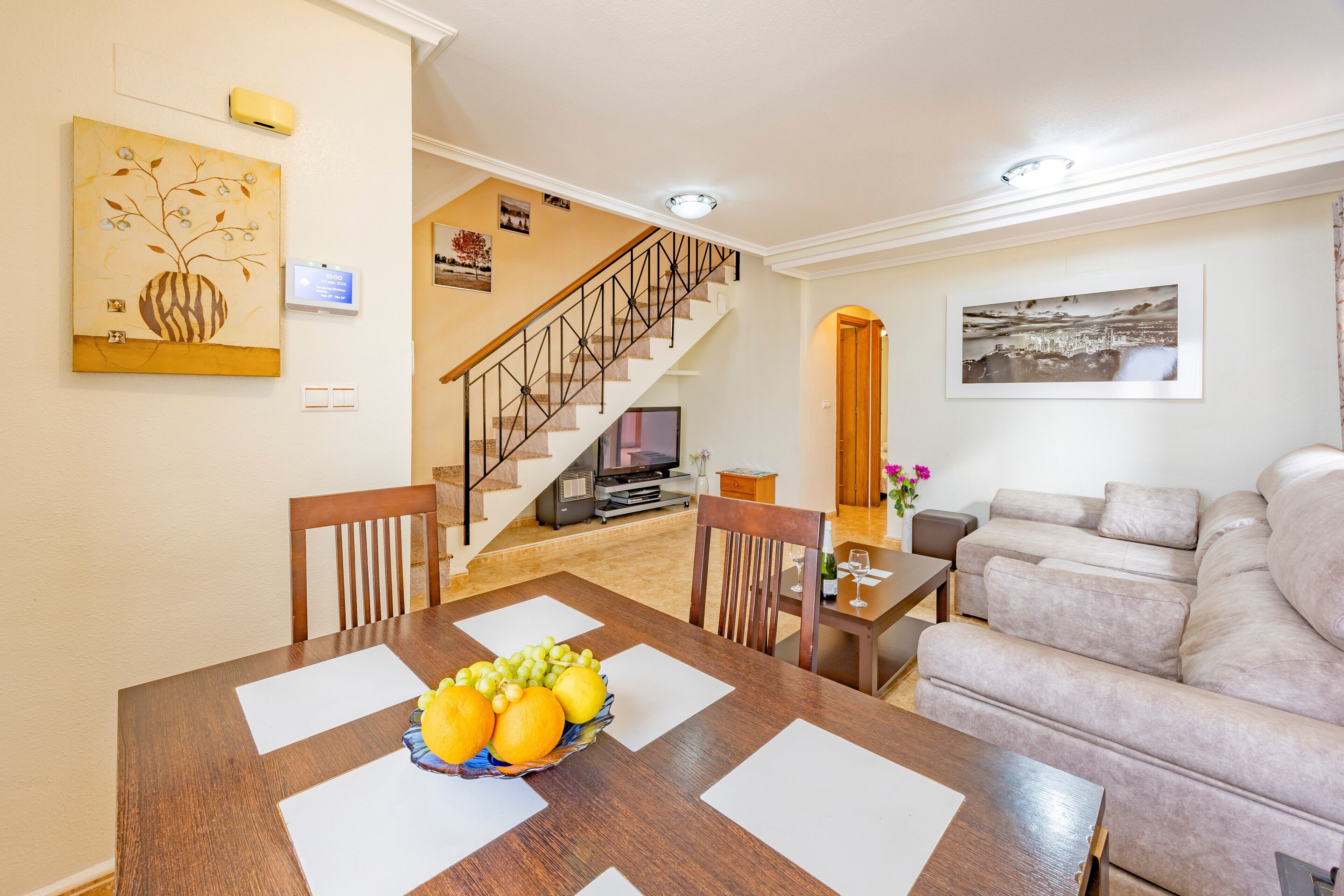 Uma sala de jantar com mesa de madeira, uma fruteira e uma escada que leva a uma sala de estar com sofá.