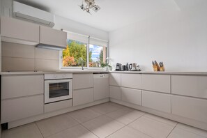 Une cuisine moderne avec des meubles blancs, un four, un évier et une fenêtre donnant sur les arbres.