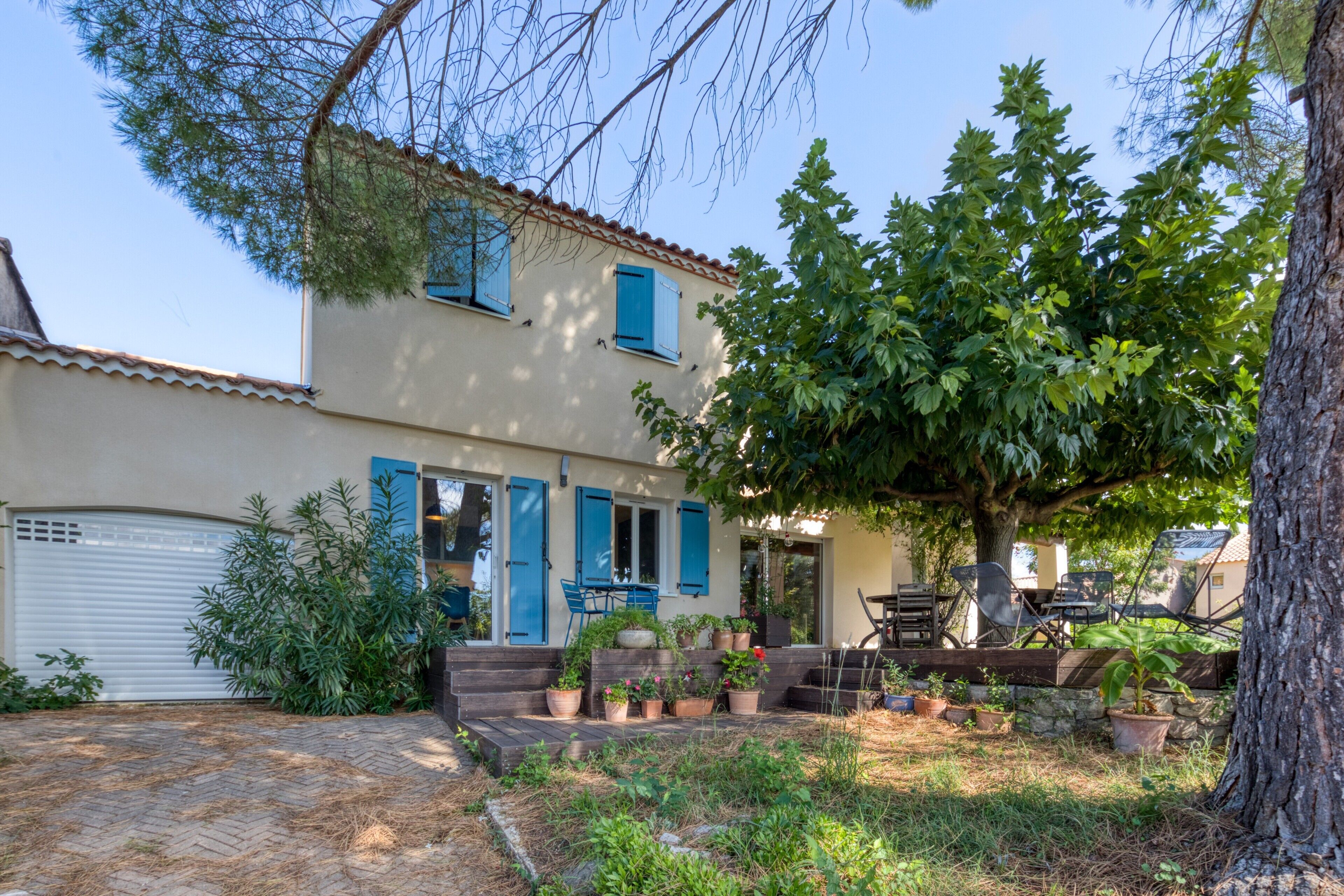 Une maison à deux étages avec un garage, une terrasse dotée de meubles d’extérieur et une variété de plantes en pot.