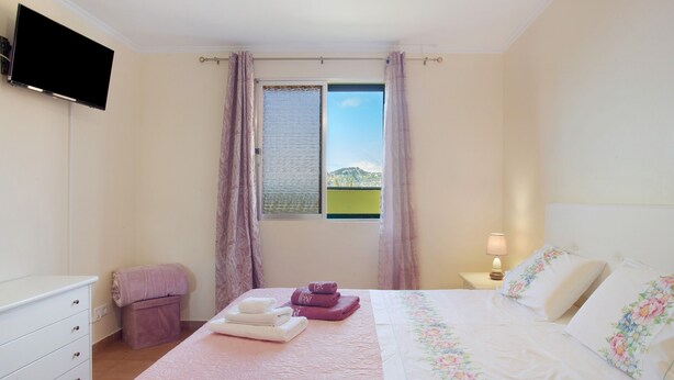 A bedroom with a bed, a dresser, a TV mounted on the wall, a window with curtains, and a nightstand with a lamp. - Holiday Home 'Levada Residence' with Private Terrace, Private Garden and Wi-Fi Access (Funchal)