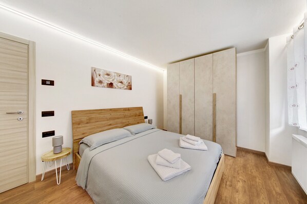 A modern bedroom with a wooden headboard, a bed with folded towels, a bedside table with a lamp, and a wardrobe.