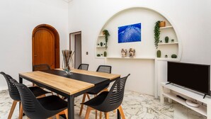 Un área de comedor con una mesa de madera y sillas negras, un televisor sobre un mueble bajo, y estantes con plantas y objetos decorativos.