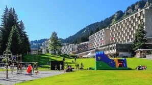 A hotel courtyard with a playground, a large building with multiple floors, and a mountainous backdrop. - Flaine Studio J2 Apartment with Mountain View and Wi-Fi (Arâches-la-Frasse)