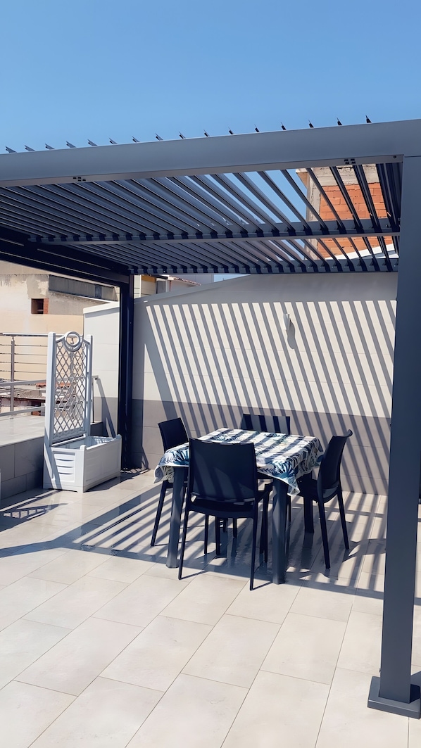 A covered outdoor patio with a table and chairs, a pergola, and a view of a building in the background.