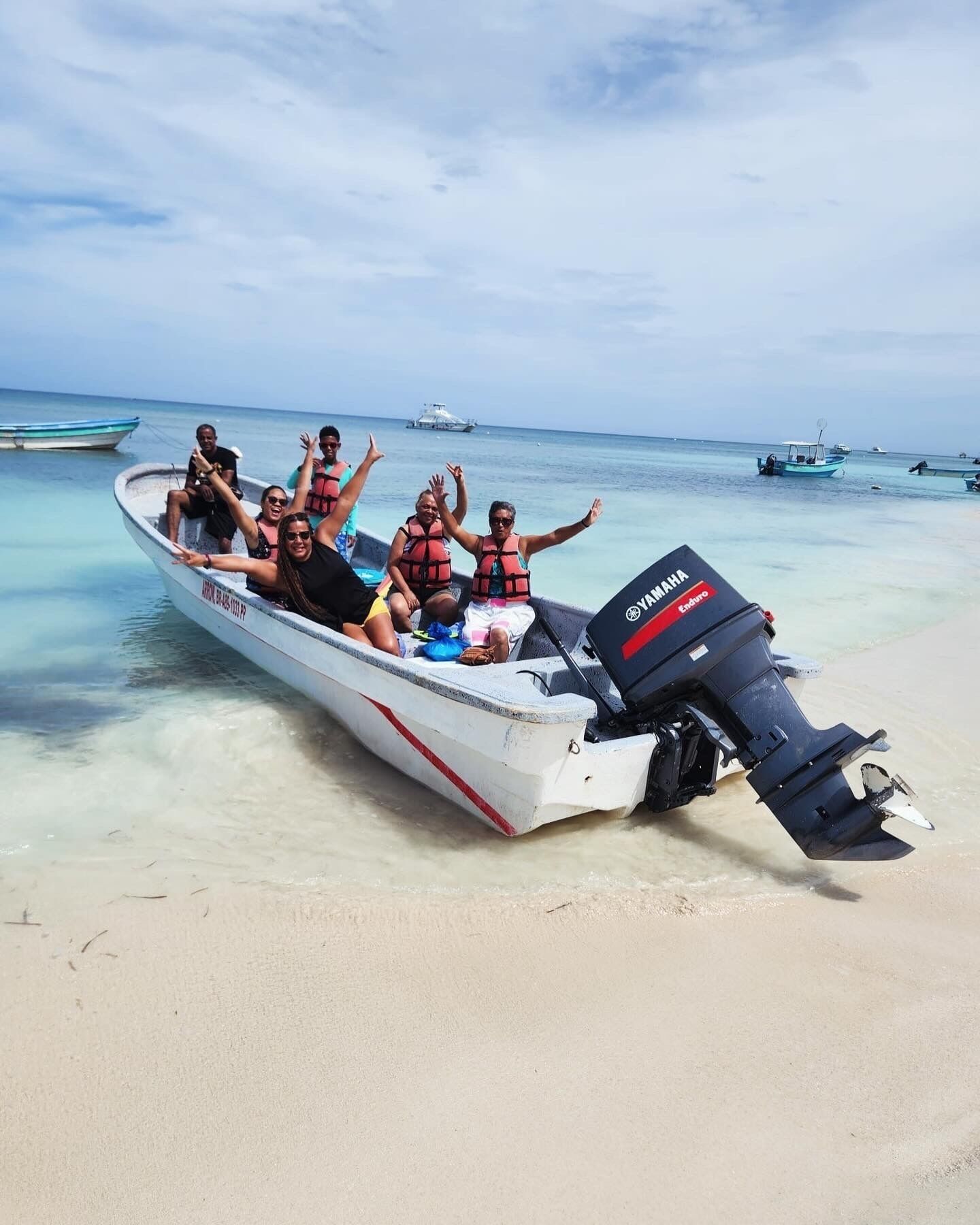 A boat with people wearing life jackets on a sandy beach with clear water and other boats in the background.