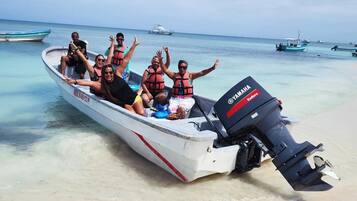 A boat with people wearing life jackets on a sandy beach with clear water and other boats in the background.