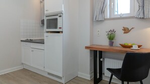 A compact kitchen with a microwave, a small refrigerator, a wooden table, a chair, and a window with checkered curtains. - Strand Loft - Ferienwohnung "strand-loft" in Westerland (Sylt)